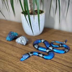 May include: A blue and silver decorative snake figurine with a detailed scale pattern. The snake is positioned on a wooden surface, near two decorative rocks and a potted plant. The snake's head is raised, and its body is coiled.