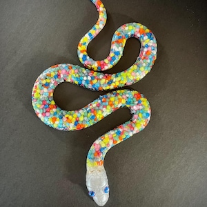 May include: A decorative snake figurine with a body covered in small, multi-coloured beads. The snake's head is silver with blue eyes. The snake is posed on a black surface.