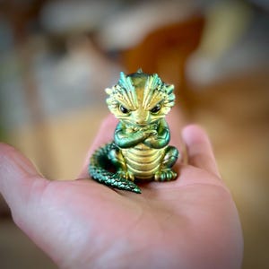 May include: A small, detailed figurine of a dragon with a gold and green iridescent finish. The dragon has its arms crossed and an angry expression. The figurine is sitting in a person's hand.