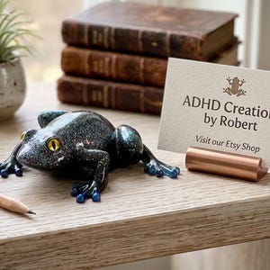 May include: A dark, glittery frog ornament with yellow eyes and blue detailing. The frog is on a wooden surface, beside a pencil and a business card that reads "ADHD Creations by Robert". Behind the frog are three antique books.