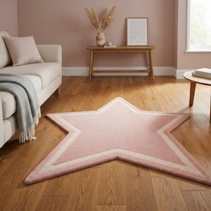 May include: A star-shaped area rug in a soft pink hue featuring a cream-coloured border detail. The rug is positioned on a wooden floor in a room with a light-coloured sofa, a wooden console table, and a small round wooden coffee table nearby.