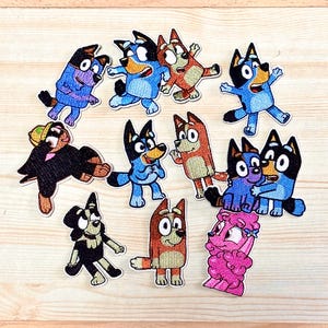 Può includere: Una collezione di toppe ricamate raffiguranti personaggi di cani dei cartoni animati in varie pose. Le toppe presentano una gamma di colori, tra cui blu, rosso, nero e rosa, con cuciture e contorni dettagliati. Le toppe sono disposte su una superficie di legno chiaro.