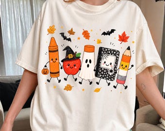 May include: An off-white t-shirt featuring a graphic print of five anthropomorphic school supplies dressed for Halloween, including a crayon, an apple with a witch hat, a ghost, a composition notebook, and a pencil, surrounded by autumn leaves and bats.