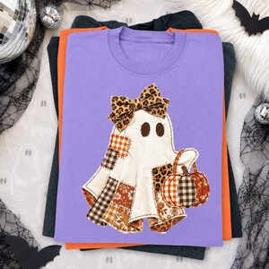 May include: A lavender t-shirt featuring a central graphic of a ghost adorned with a leopard print bow. The ghost is composed of various patchwork fabric patterns in orange, brown, and white, and it is holding a small pumpkin with a matching pattern.