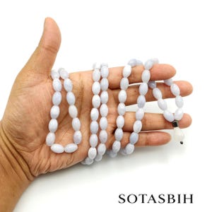 May include: A string of light blue, oval-shaped beads arranged in multiple loops. The beads have a smooth, semi-translucent finish. A single, white, teardrop-shaped pendant is attached to one end with a small dark metal spacer. Text reads SOTASBIH.
