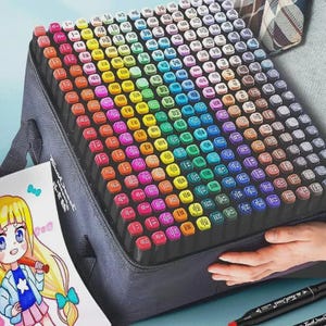 May include: A grey art case packed with a multitude of coloured markers, arranged in a rainbow spectrum. A drawing of an anime character is in the foreground. A black marker is visible in the bottom right corner.