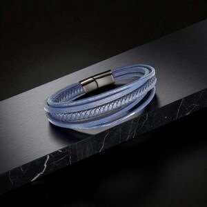 May include: A light blue leather bracelet with multiple strands and a braided detail. The bracelet has a silver-tone magnetic clasp. The bracelet is displayed on a dark, textured surface.