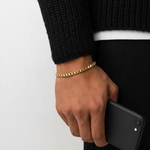 以下が含まれることがあります： 長方形のリンクデザインのゴールドカラーのブレスレット。ブレスレットは人の手首に着用されています。また、その人は黒いスマートフォンを持っています。背景は無地です。