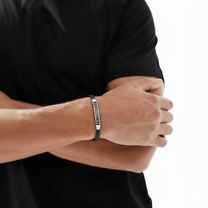 May include: Black leather bracelet with a silver-tone rectangular centerpiece. The centerpiece features a geometric design with black accents. The bracelet is worn on a person's wrist, with the arm and torso visible.