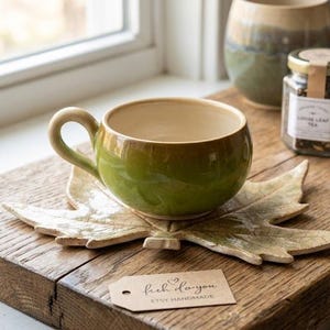 Puede incluir: Una taza de té de cerámica hecha a mano con exterior verde e interior beige, sobre un platillo en forma de hoja. La taza está sobre una superficie de madera, junto a un tarro de té y un paño enrollado. La etiqueta dice "fish do you, etsy handmade."