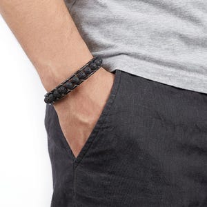 May include: A black lava stone bracelet with two rows of dark, porous beads. The bracelet is woven with a dark cord and features a silver-coloured clasp. The bracelet is worn on a wrist, with the person wearing a grey t-shirt and dark grey trousers.