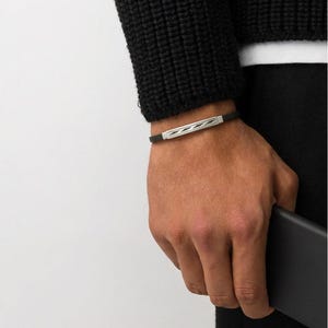 May include: A black leather bracelet with a silver bar featuring a geometric design. The bracelet is worn on a wrist, with a black sweater and pants in the background. The person is holding a black phone.