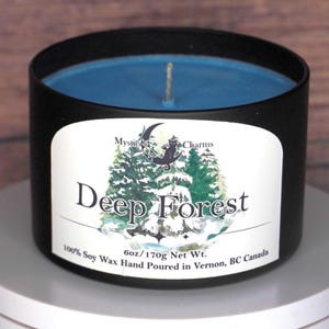 May include: A black metal candle tin containing blue soy wax with a single wick. The label features a crescent moon, a cat, and a forest illustration with the text Mystic Charms, Deep Forest, and 6oz/170g Net Wt. 100% Soy Wax Hand Poured in Vernon, BC Canada.