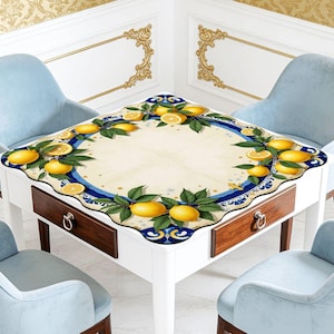 K&ouml;nnte beinhalten: Ein wei&szlig;er quadratischer Tisch mit zwei Schubladen und einer Tischdecke mit Zitronenmotiv, umgeben von vier hellblauen St&uuml;hlen mit Holzbeinen. Die Tischdecke hat einen Rand aus Zitronen und gr&uuml;nen Bl&auml;ttern.