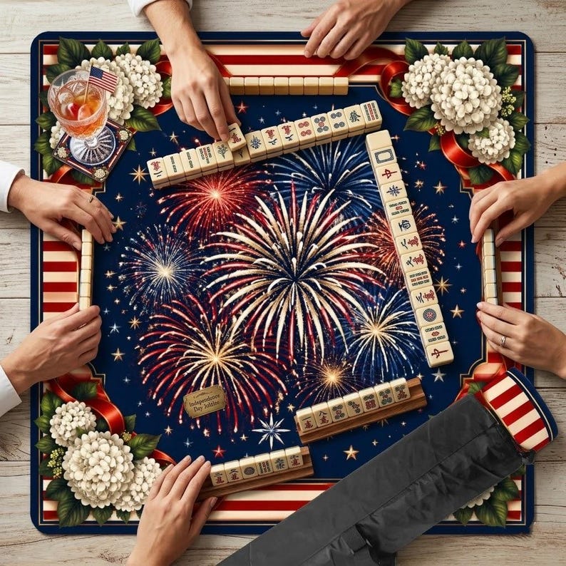 May include: A Mahjong game in progress on a navy blue mat with a fireworks design. The mat has red and white stripes around the edges and white floral accents. Mahjong tiles are arranged around the mat, with hands holding the tiles. A cocktail with an American flag is on the mat.