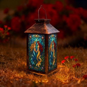 May include: A rectangular lantern with a dark, weathered bronze-coloured frame and a wire handle. The four glass panels feature a stained-glass style illustration of two mermaids with long flowing hair in blue and gold tones, set against a blue grid.
