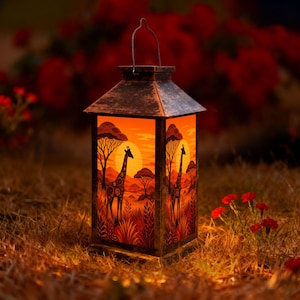 May include: A rectangular lantern with a distressed dark metal finish and a wire handle. The side panels feature a vibrant orange and yellow sunset illustration depicting giraffes, acacia trees, and savanna foliage, illuminated from within.