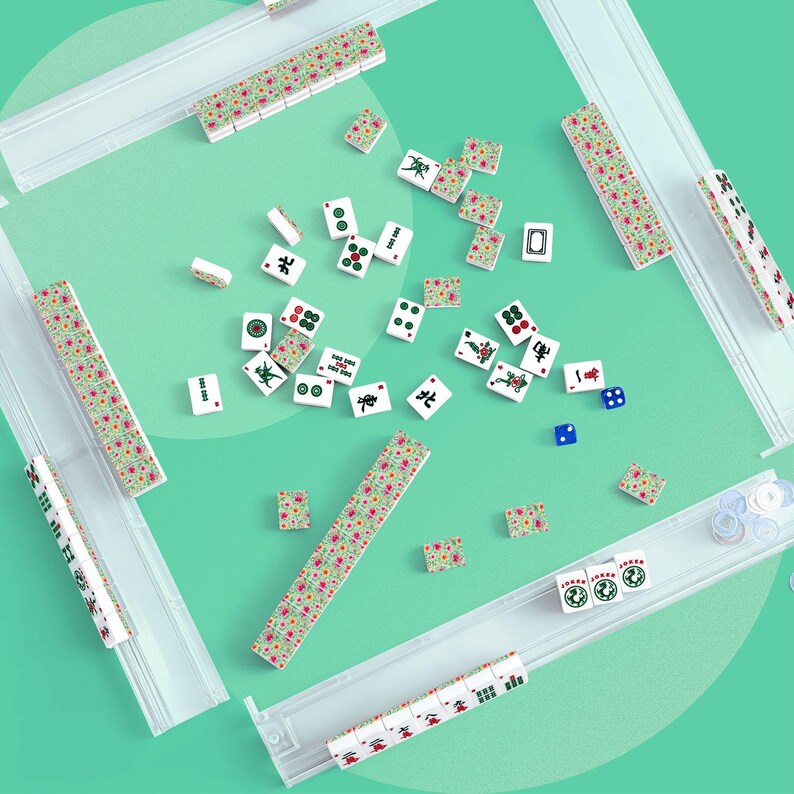 K&ouml;nnte beinhalten: Ein Mahjong-Spielset mit wei&szlig;en Spielsteinen, die florale Muster auf der R&uuml;ckseite sowie gr&uuml;ne, rote und schwarze Schriftzeichen und Zahlensymbole aufweisen. Das Set enth&auml;lt vier transparente Kunststoffhalterungen, zwei blaue W&uuml;rfel und Spielmarken.