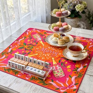 K&ouml;nnte beinhalten: Ein pink-oranges Tischarrangement mit Mahjong-Spiel, mehrst&ouml;ckigem Dessertst&auml;nder mit Macarons und Scones sowie einer Teetasse. Auf dem Tischl&auml;ufer steht das Wort "Marry".
