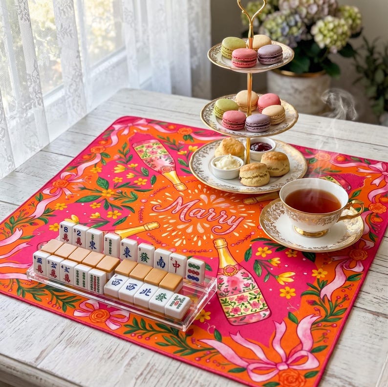 K&ouml;nnte beinhalten: Ein pink-oranges Tischarrangement mit Mahjong-Spiel, mehrst&ouml;ckigem Dessertst&auml;nder mit Macarons und Scones sowie einer Teetasse. Auf dem Tischl&auml;ufer steht das Wort "Marry".