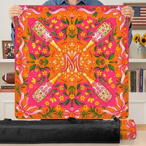May include: A square, colorful tablecloth with a vibrant design. The tablecloth features champagne bottles, floral patterns, and the letter "M" in the center. The color scheme includes pink, orange, and yellow, with ribbon and bow accents. The tablecloth is approximately 40 inches square.