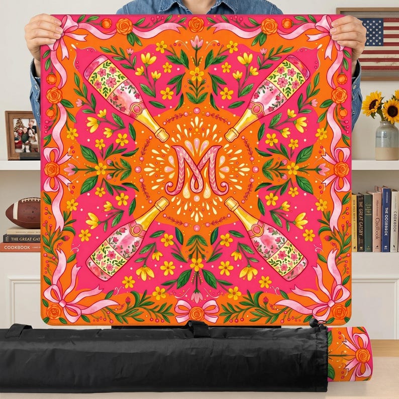 May include: A square, colorful tablecloth with a vibrant design. The tablecloth features champagne bottles, floral patterns, and the letter "M" in the center. The color scheme includes pink, orange, and yellow, with ribbon and bow accents. The tablecloth is approximately 40 inches square.