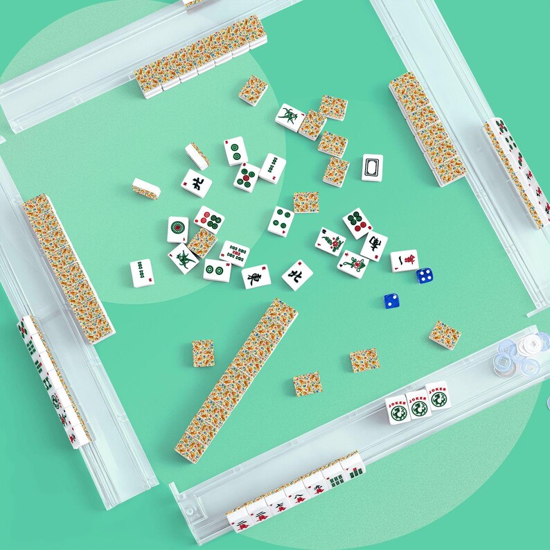 May include: A Mahjong set featuring white tiles with colourful character, bamboo, and dot designs, along with patterned backs. The set includes four clear plastic tile racks, two blue dice, and several circular white tokens arranged on a green surface.