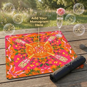 K&ouml;nnte beinhalten: Eine leuchtend rosa-orangefarbene Picknickdecke mit Blumen- und Champagnerflaschen-Design. Die Decke hat eine aufgerollte schwarze Tragetasche. Auf der Decke steht "Add Your Monogram Here". Ein Glas Champagner mit einer rosa Rose steht auf dem Holztisch.