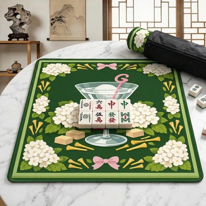 Peut inclure: Tapis de Mahjong vert avec un motif floral, un verre &agrave; cocktail et des tuiles de Mahjong. Le tapis pr&eacute;sente des fleurs blanches, des n&oelig;uds roses et des tees de golf jaunes. Un &eacute;tui de transport noir est visible.