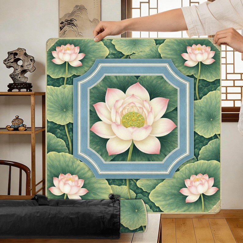 May include: A square, floral-themed mat featuring a central lotus flower with pink and white petals, surrounded by green leaves. The design includes a light blue frame and four additional lotus flowers. The mat is held up by a person.