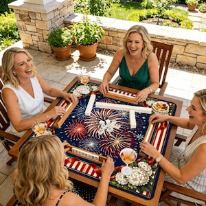 May include: A wooden mahjong table with a navy blue surface featuring a fireworks design in red, white, and gold. Four women are seated around the table, playing mahjong. The table is set outdoors on a patio.