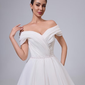 Peut inclure: Robe de mariée blanche à épaules dénudées avec un décolleté en cœur. Le corsage présente un design drapé et la taille est ornée d'une ceinture perlée. La jupe est ample et fluide, créant une silhouette élégante.