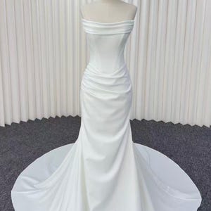 Può includere: Un abito da sposa bianco senza spalline su un manichino. L'abito presenta un corpetto aderente con sottili arricciature e una silhouette a sirena che si allarga in un lungo strascico. Il tessuto sembra essere un raso liscio.
