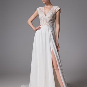May include: A white wedding dress with a V-neck lace bodice and a flowing skirt with a high slit. The dress has cap sleeves and a subtle train. The model is wearing white sandals. The dress is elegant and suitable for a wedding.