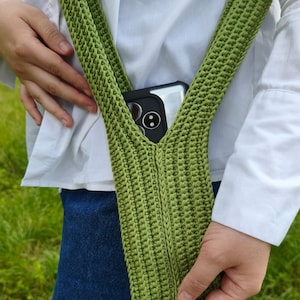 May include: A sage green crocheted crossbody phone pouch featuring a textured vertical stitch pattern and a wide integrated shoulder strap. The pouch contains a black smartphone with a visible camera lens array, showcasing a functional design.