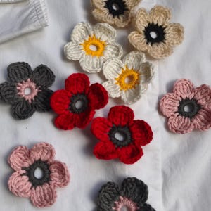 Peut inclure: Une collection de dix fleurs crochetées à la main dans diverses couleurs, notamment rouge, rose, blanc, beige et gris foncé. Chaque fleur présente cinq pétales arrondis et un centre circulaire dans une couleur de fil contrastante sur un tissu blanc.