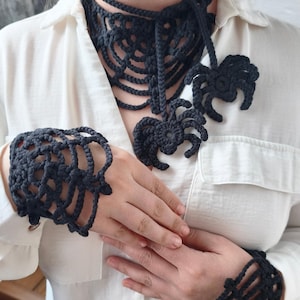 May include: A black crocheted choker necklace featuring a spiderweb pattern and two dangling spider motifs, paired with matching black crocheted spiderweb fingerless gloves. The set is worn over a plain white button-down shirt with a collar.