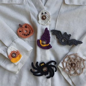 Peut inclure: Un ensemble de six couvre-boutons crochetés sur le thème d'Halloween comprenant un fantôme blanc, une citrouille orange, un chapeau de sorcière violet, une chauve-souris grise, un bonbon jaune et orange et une araignée noire.