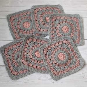 May include: A collection of five square crochet motifs featuring a central dusty pink circular pattern surrounded by grey yarn. Each piece is crafted with a textured stitch design, forming a floral-like centre that transitions into a square border.