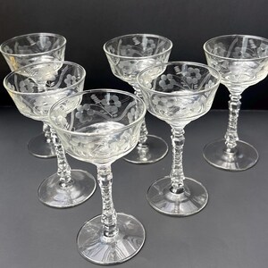 May include: A set of six clear glass coupes featuring etched floral patterns on the bowls. Each glass has a multi-faceted, textured stem and a circular base. The design includes delicate lines and small flower motifs etched into the glass surface.