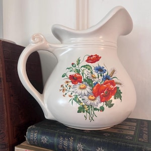 May include: A cream-colored ceramic pitcher featuring a curved handle and a wide pouring spout. The front of the pitcher is decorated with a colorful floral illustration of red poppies, white daisies, blue cornflowers, and stalks of wheat.