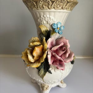 May include: Cream-colored ceramic vase with a scalloped rim and ornate detailing. The vase features three-dimensional, colorful floral accents, including yellow, pink, and blue flowers. The vase is supported by three small feet.