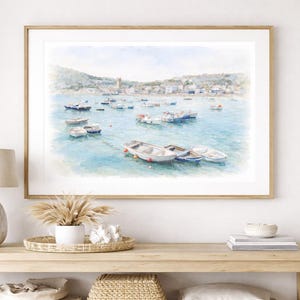 May include: Watercolor print of a coastal scene with numerous small boats floating on turquoise water. The artwork, framed in a light wood, depicts a quaint village nestled along the shoreline under a soft blue sky.