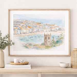 May include: Watercolor print of a coastal town with buildings, boats, and a church tower. The artwork is framed in a light wood frame. The scene is painted in soft blues, greens, and beige tones. The print is displayed on a light wood table with decorative items.
