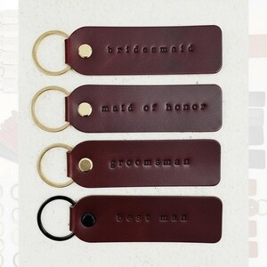 May include: Four burgundy leather keyrings with gold and black hardware. Each keyring is rectangular with rounded edges and embossed text: "bridesmaid", "maid of honor", "groomsman", and "best man".