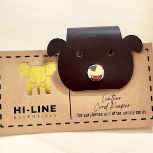 May include: A dark brown leather cord keeper shaped like a bear's head, featuring a gold button nose. It's attached to a brown card with the text "HI-LINE ESSENTIALS" and "Leather Cord Keeper for earphones and other unruly cords."