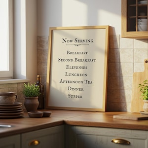May include: A framed sign with the text "Now Serving" and a list of meals, including breakfast, lunch, and dinner, is displayed in a kitchen setting. The sign is in a light wood frame. A stack of plates and a cup are on the counter.