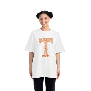 Puede incluir: Una camiseta blanca con una gran "T" naranja compuesta por palabras como "Rocky Top" y "Tennessee". La persona que la lleva, con el pelo oscuro recogido, sonríe. La camiseta tiene un corte holgado y de estilo informal.