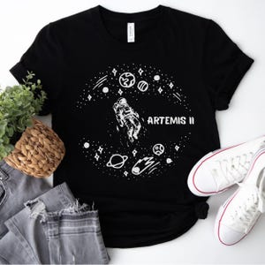 Może przedstawiać: Czarny t-shirt z białą grafiką przedstawiającą astronautę w kosmosie, otoczonego planetami, gwiazdami i napisem "ARTEMIS II". Koszulka jest zestawiona z szarymi dżinsami i białymi trampkami.