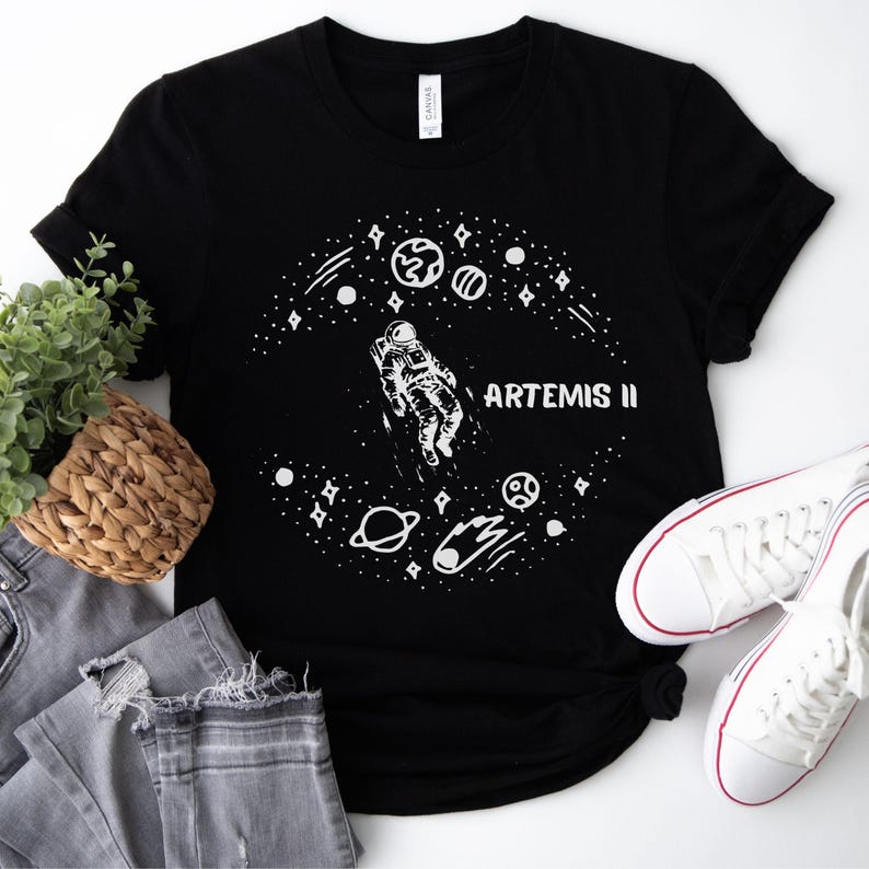 Może przedstawiać: Czarny t-shirt z białą grafiką przedstawiającą astronautę w kosmosie, otoczonego planetami, gwiazdami i napisem "ARTEMIS II". Koszulka jest zestawiona z szarymi dżinsami i białymi trampkami.